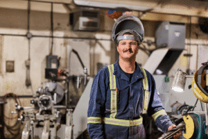 a smiling man standing standing by machinery. He is wearing safety glasses, welder's hat, blue coveralls, and a yellow high-visibility vest.